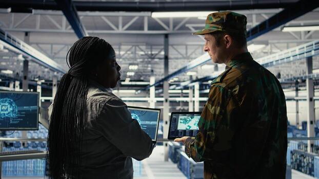 Military commander and female programmer in data center tracking coordinates using notebook software. Soldier and african american woman in server hub analyzing reconnaissance data, camera A photo
