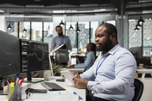 Trader at work tracking stock exchange metrics and placing orders using trading software on PC. African american man in brokerage company office trading securities on behalf of clients photo