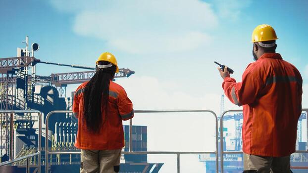 Offshore platform engineer reports operation status using walkie talkie, following safety protocols. African american man in charge of drilling rig communications doing checkup on equipment, photo