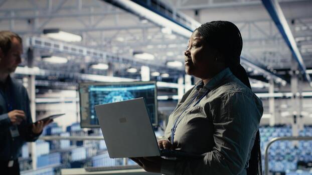 Data center supervisor on industrial platform working to improve AI rigs processing speed. African american server hub manager overseeing gear supporting artificial intelligence workloads, camera B photo