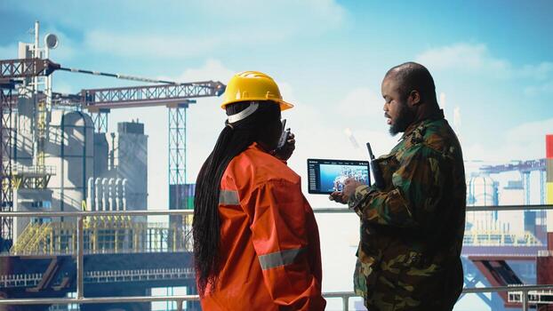 Soldier on offshore platform deck talking with technician, using device visualization software. African american naval unit guards drilling rig against terrorist threats using tablet app, photo
