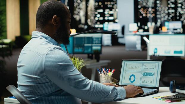 Black advisor researching market data and building strategy for business targets, using spreadsheets on laptop for proofreading. Late night overtime for a multinational company. photo
