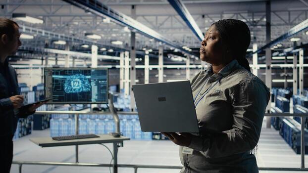 Data center admin monitors deep learning process with laptop, reviewing operational metrics. African american woman using device to review neural networks reports during inspection, camera A photo