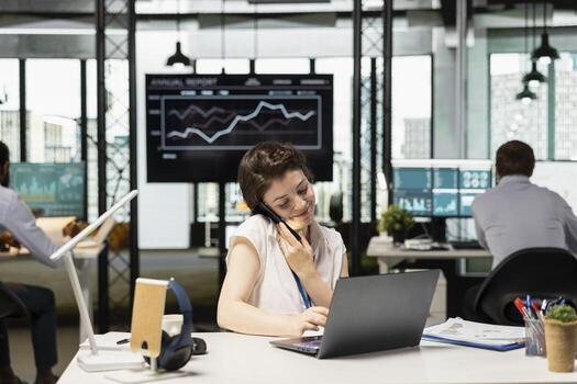 sonriente caucásico oficina trabajador multitareas, hablando en móvil teléfono y utilizando ordenador portátil a escritorio. hembra empleado en llamada con colega, lluvia de ideas negocio soluciones en moderno lugar de trabajo. foto