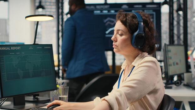 Young businesswoman focused on a podcast while she resolves office tasks, monitoring insights or KPI metrics on pc. Strategy reports for expansion plans with digital tools. Camera B. photo