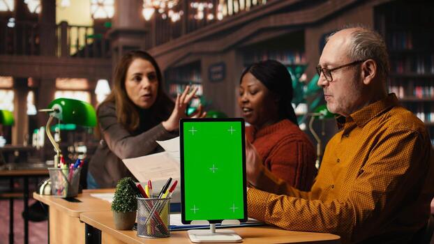 Mature colleagues team review paperwork next to chroma key mockup, using multiple academic sources and books in classic library setting. Learning space with library resources. photo