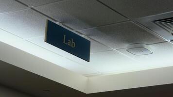 Ceiling interior view of a building hallway, featuring a rectangular sign with white text that reads - Lab. Medical, scientific, or educational laboratory environment video