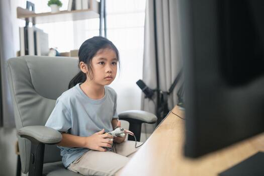 Young girl focused on game console while sitting in a comfortable chair in a bright room with modern decor and computer setup photo