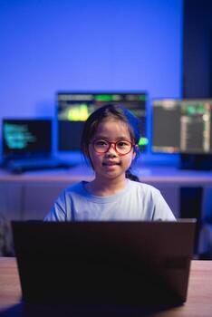 Young girl with glasses working on a laptop in a tech-savvy workspace with multiple computer screens displaying data and programming codes, focused expression. photo