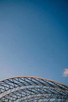 Vertical screen. curved metal beams creating robust and intricate framework rise against clear blue sky, highlighting elegance of industrial design and contemporary architecture. Modern architecture photo