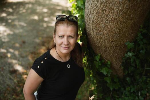 de edad mediana mujer sonriente y descansando, ganando fuerza después un trabajando día en naturaleza. confidente mujer propensión en contra un árbol cubierto en hiedra, con sereno natural antecedentes foto