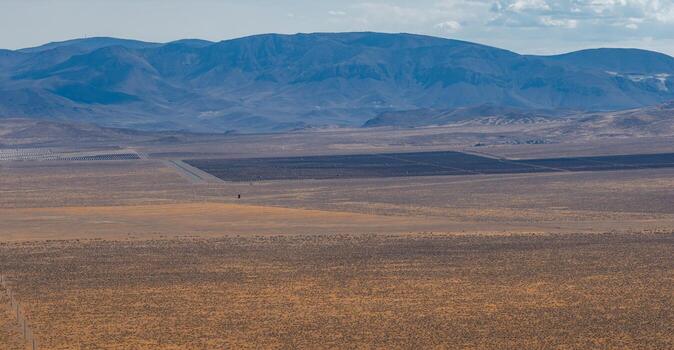 Expansive desert terrain in Nevada with dark rectangular industrial sections, sparse vegetation, and rugged mountains under a partly cloudy sky. photo