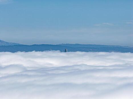The tip of Salesforce Tower rises above a dense cloud layer, with faint hills in the background under a clear blue sky in San Francisco. photo