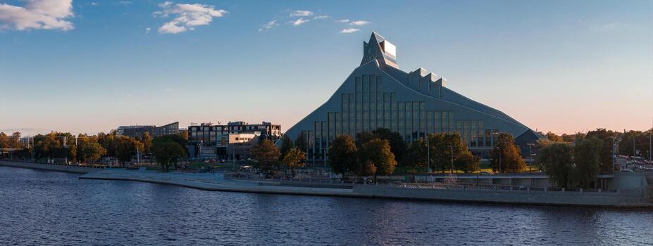 The National Library of Latvia with its pyramid like design reflects the sky, set along the Daugava River with trees and a promenade in Riga. photo