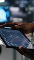 Vertical Close up of data center engineer using tablet to review code for automation scripts. Server farm system administrator using device, monitoring error logs for debugging processes, camera A video