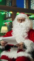 vertikal porträtt av man spelar jul karaktär läsning brev från ungar, hyssande publik med finger gest. santa claus be för stillhet medan läsning barn önskar listor, kamera en video