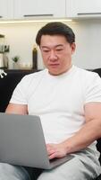 asiático hombre a hogar lanza ordenador portátil botas sistema y comienza trabajo mecanografía con calma sonrisa. chino chico en sofá abre aplicación cheques bandeja de entrada conjuntos plan para día y empieza primero tarea luego conjuntos Temporizador para atención video