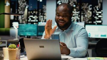 Black employee chatting late at night on phone web teleconference, engaged in a digital discussion for productivity. Interacting with shareholders. Networking session in corporation. Camera B. video