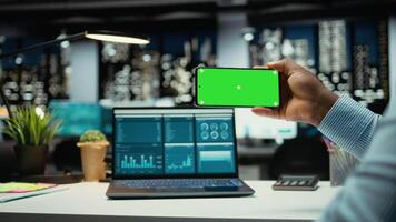Preto escritório empregado trabalhando com isolado tela em Móvel telefone, revisão e facto verificação dentro a escritório atrasado às noite. pessoa colecionar em formação em uma computador portátil para por mês relatório. Câmera uma. video