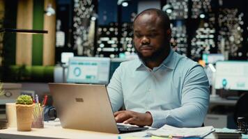 Afrikaanse Amerikaans Mens doet kritiek denken en proeflezen met papieren voordat transcriberen gegevens Aan notitieboekje, zittend in de kantoor Bij nacht. arbeider documenteren belangrijk ideeën. camera b. video