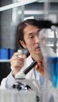 Vertical Multiethnic nanotechnology researchers using microscope to evaluate cellular structures in lab. Diverse workers observes medical tissue through magnification tool at laboratory bench, camera B video