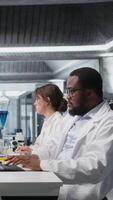 Vertical Quality control analyst in lab testing blood sample using computer. African american man in laboratory analyzing sanguine fluid specimen in vial used for medical research with PC software, camera A video