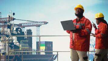 afrikansk amerikan arbetstagare på offshore plattform däck konfigurera borrtorn förvaltning system för Mer effektiv olja extraktion. borrning rigg fartyg expert- håller på med havsbotten analys använder sig av bärbar dator, kamera en video