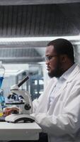 verticale laboratorio scienziato utilizzando microscopio per valutare cellulare strutture. africano americano uomo osserva campioni attraverso ingrandimento attrezzo a laboratorio panca, avanzando scientifico conoscenza, telecamera un' video