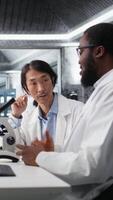 vertikal team av multietnisk forskare använder sig av mikroskop till utvärdera cellulär strukturer i labb. raser kollegor observera medicinsk vävnad genom förstoring verktyg på laboratorium bänk, kamera en video