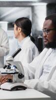 vertikal forskare rörelse mikroskop, håller på med prov observationer i laboratorium, skrivning anteckningar. forskare studier vävnad under optisk enhet i bioteknik labb, rapportering fynd, kamera b video
