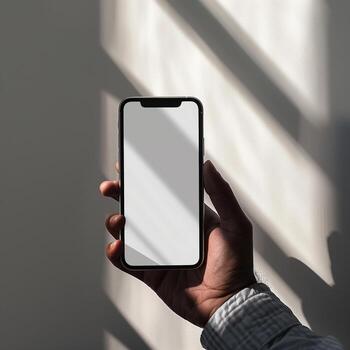 Person holding a modern smartphone with an empty display against a textured wall with window light and shadows. photo