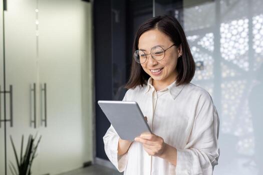 Young beautiful Asian woman with tablet computer standing near window, smiling businesswoman using online application, programmer testing new software. photo