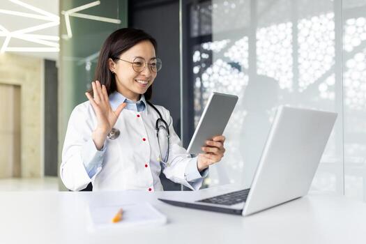 Young Asian female doctor working inside medical office consulting patients remotely, smiling with satisfaction at client, using tablet computer for call, sitting at desk with laptop. photo