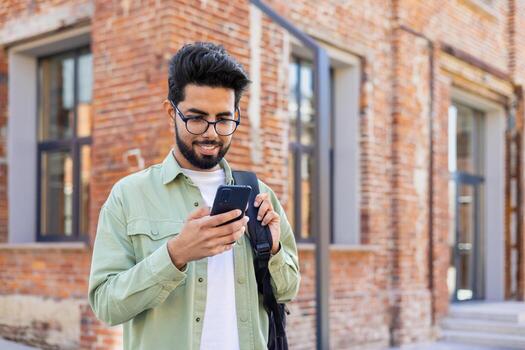 Portrait of young successful student outside university campus, man smiling and using app on phone, typing message and browsing social media, indian man holding phone with backpack. photo