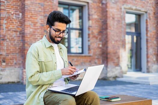 Young man studying remotely sitting on a bench outside a university building, a student in headphones watching a online course, talking using a call, writing down data in a notebook. photo