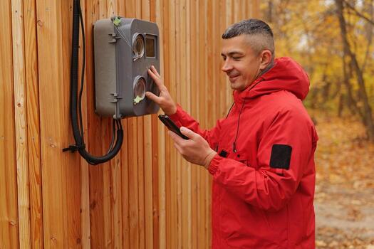 A man wearing a red jacket smiles as he checks his phone while standing near a device mounted on a wooden structure in a forest setting photo