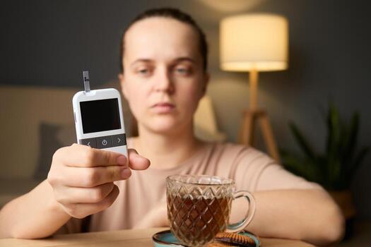 A person is checking their blood sugar levels using a glucometer while sitting at a table with a cup of tea and cookies. Soft lighting creates a warm atmosphere photo