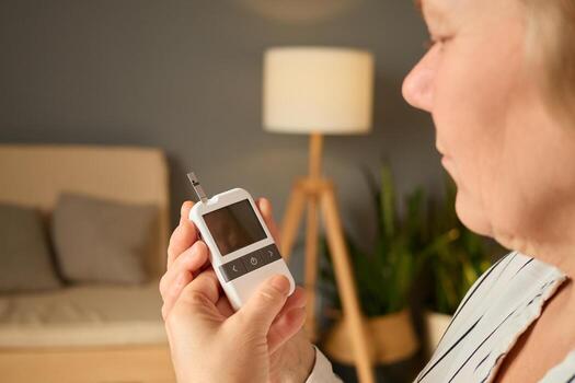 A person checks their blood sugar levels using a glucometer while seated in a comfortable living room. The atmosphere is warm and inviting, with a lamp and plants in the background photo