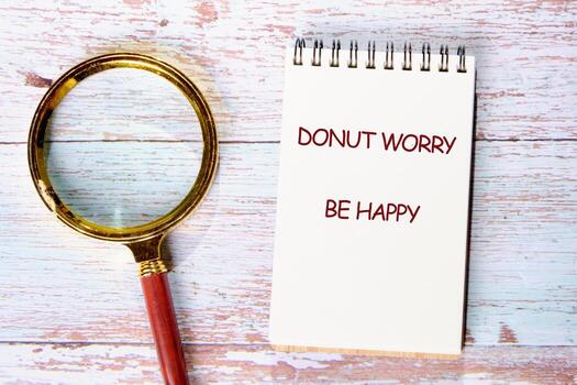 A spiral notebook sits on a wooden surface with the phrase Donut Worry Be Happy written on it. Next to the notebook is a magnifying glass, perhaps searching for joy photo
