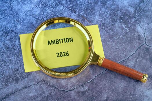 A yellow note with the word Ambition and the year 2026 is highlighted by a gold-rimmed magnifying glass on a textured background. Future planning, goal setting, and success are implied photo