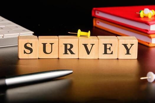 Wooden blocks spell out SURVEY. The blocks are arranged on a desk near books, a pen, a keyboard, and colorful pushpins. The surface reflects the light photo