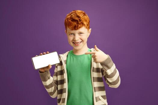 Isolated Portrait of Joyful Red Haired Child Boy Holding Smartphone and Showing to Camera Empty Screen of it Display, Points There With her Finger, Smiles Standing Against Violet Isolated Background photo