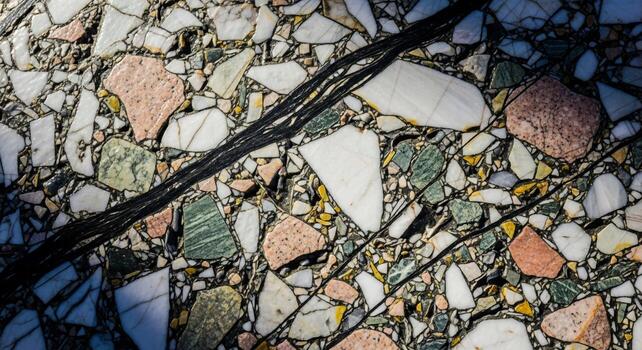 Close up abstract view of a dark rough textured object diagonally crossing a surface composed of multi colored aggregate stone fragments photo