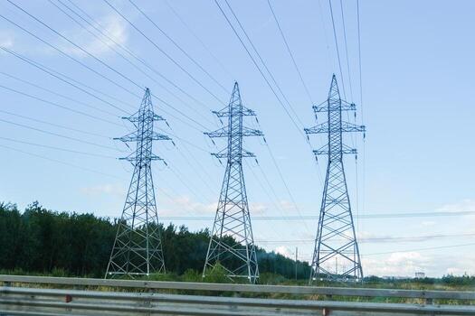Electrical power lines extending between multiple high-voltage pylons against a light blue sky with trees in the background, symbolizing energy transmission. photo
