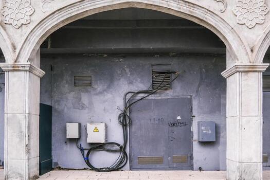 fragmento de urbano edificio demostración arqueado columnas enmarcado un desgastado pared con enredado eléctrico cable, utilidad caja, y cerrado metal puerta con graff foto