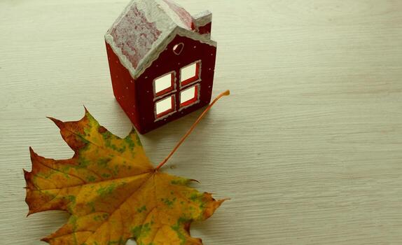 A light oak shelf with a fallen maple leaf and a miniature house copy space image. A stock photo of autumn symbols and a cozy home with empty space at a side