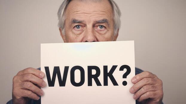 Senior Caucasian man holding a sign that reads WORK with a concerned expression. The background is neutral and simple, photo
