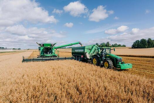 A green combine harvester transfers grain to a tractor's cart in a vast golden field, with rows of crops and a tree line on the horizon under a cloudy sky. photo
