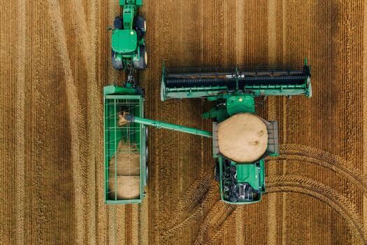 An aerial view shows a green combine harvester unloading grain into a green tractor with a trailer on a golden brown field with visible tire tracks. photo