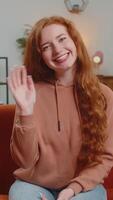 contento joven caucásico mujer sonriente simpático a cámara y ondulación manos gesticulando Hola, Hola, saludo hablando mientras sentado en sofá en vivo habitación a hogar. contento pelirrojo niña grabación vlog a hogar. video
